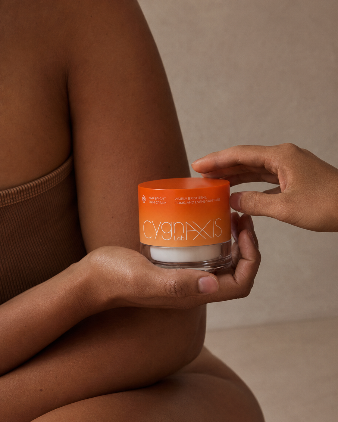 Hands holding NVP Bright Firm Cream against a warm neutral background.