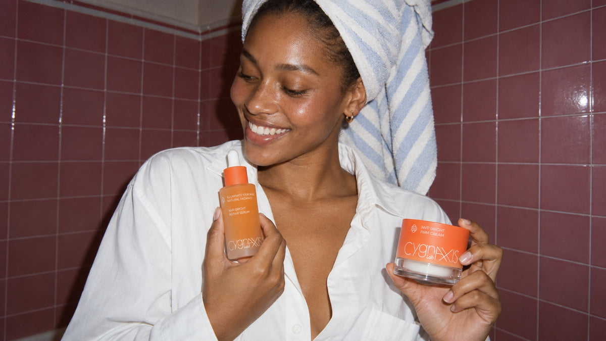 Woman holding NVP Bright Repair Serum and NVP Bright Firm Cream in a bathroom.